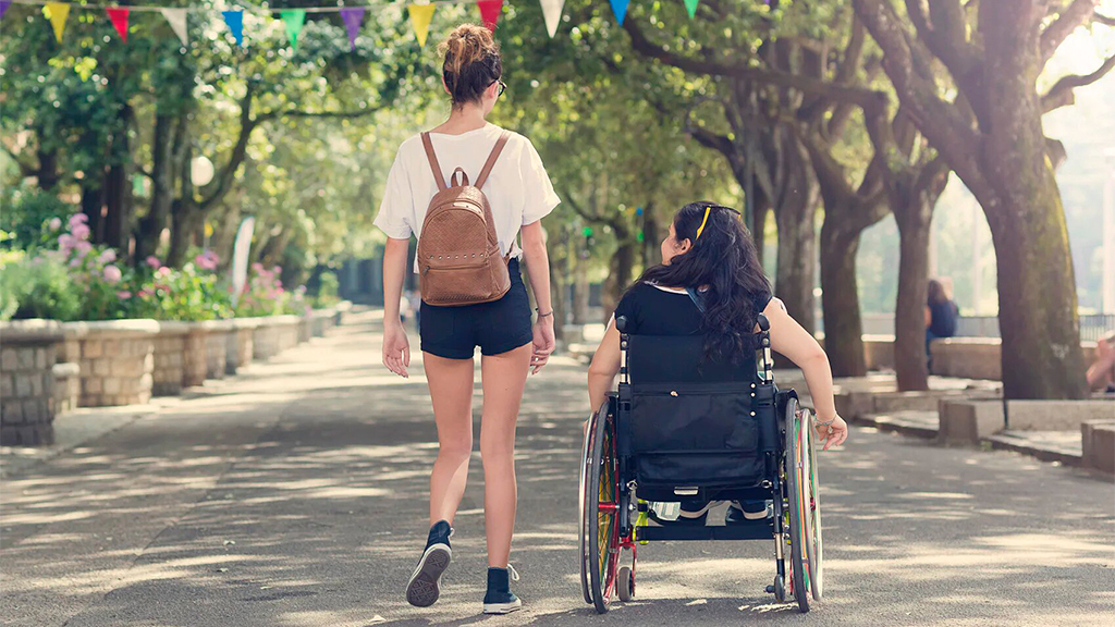 Dos mujeres jóvenes,una en silla de ruedas, paseando por un parque